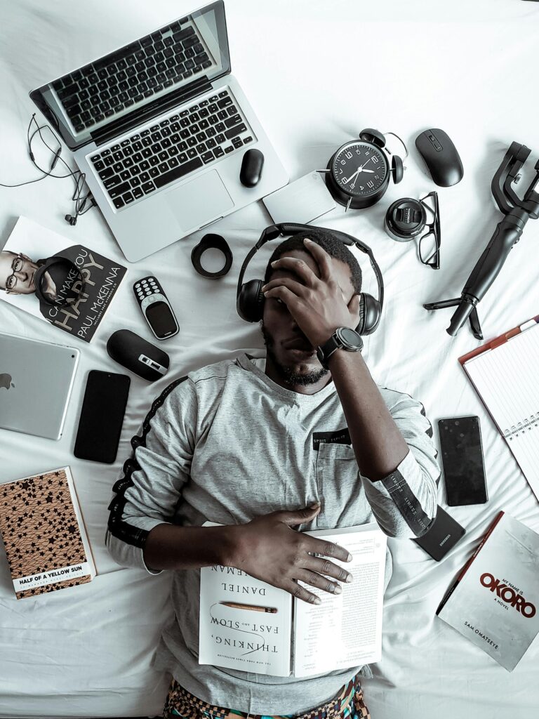 pexels-photo-3345882-3345882 A young man lying on a bed surrounded by modern gadgets, books, and tech devices, illustrating digital lifestyle.