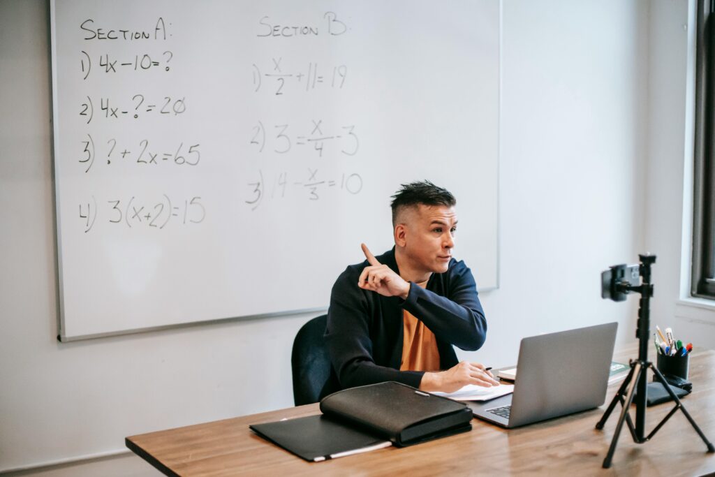 A male teacher explains math equations during an online class, using a laptop and whiteboard.