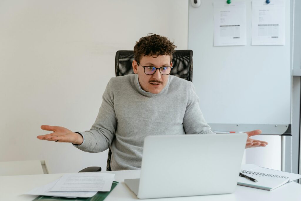 A man in a gray sweater and glasses gesturing at his laptop screen during a video call in an office setting.