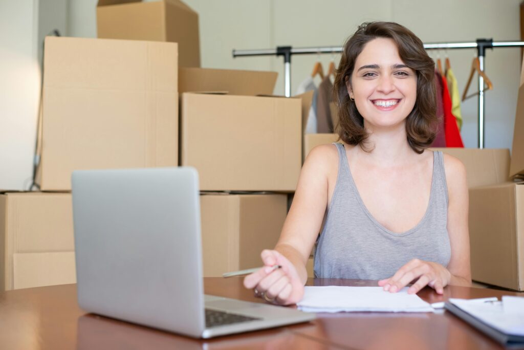 pexels-photo-7857587-7857587 Happy woman working on a laptop surrounded by cardboard boxes. Ideal for business and online store concepts.