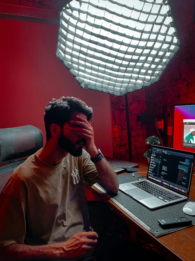 A tired man sits at a cluttered desk with a laptop under softbox lighting, looking stressed.